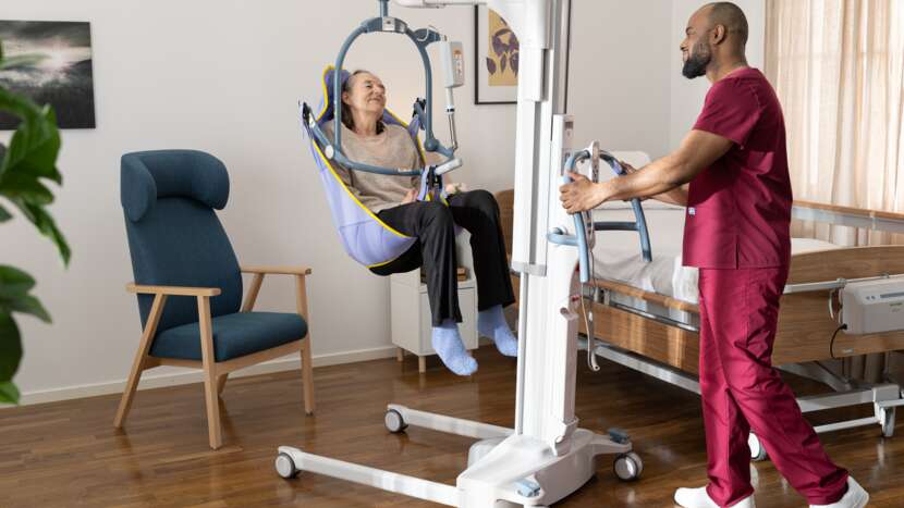 Arjo Maxi Move 5 Multifunctional Powered Patient Floor Lift With Patient And Caregiver In Bed To Chair View