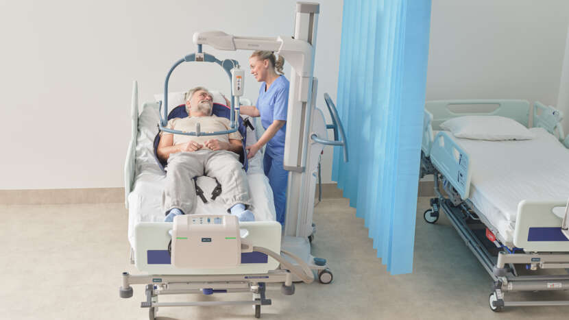 Arjo Maxi Move 5 Multifunctional Powered Patient Floor Lift With Patient And Caregiver In Bed Transfer View