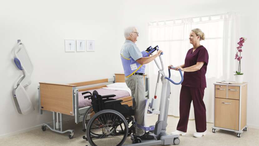 Arjo Sara Flex Sit to Stand Lift With Patient And Caregiver In Wheelchair To Bed Transfer View