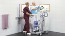 Arjo Sara Flex Sit to Stand Lift With Patient Standing Holding Handlebar And Caregiver Assisting In Bathroom View