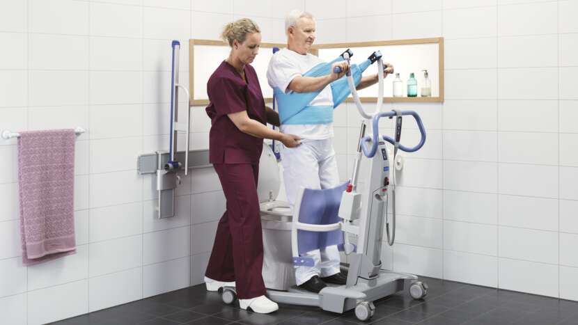 Arjo Sara Flex Sit to Stand Lift With Patient Standing Holding Handlebar And Caregiver Assisting In Bathroom View