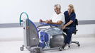 Arjo Sara Plus Sit to Stand With Patient And Caregiver Holding Hand Control Assisting Patient In Rehabilitation View