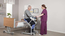 Arjo Sara Stedy Compact With Patient Sitting In And Caregiver Assisting Patient In Bed Transfer View