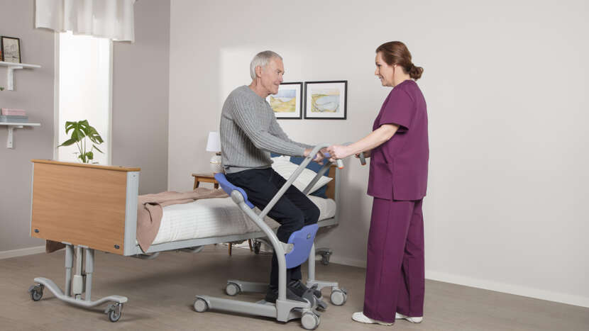 Arjo Sara Stedy Compact With Patient Sitting In And Caregiver Assisting Patient In Bed Transfer View