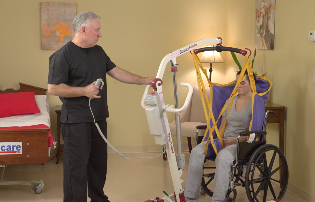 Bestcare BestLift PL400EF Foldable Patient Lift With Caregiver Holding the Hand Control And Patient Being Lifted In A Sling