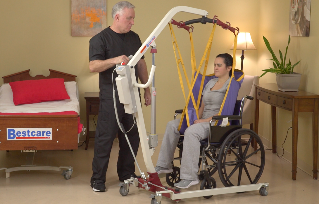 Bestcare Bestlift Full Body Lift PL182 lifting model patient in sling over wheelchair with caretaker holding hand control.