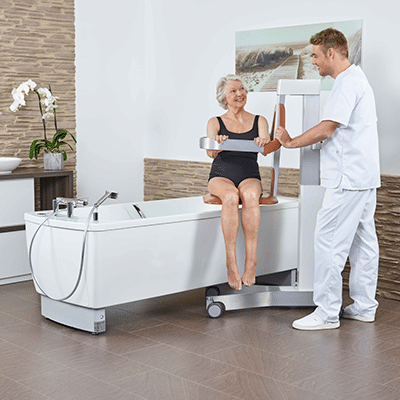 Handicare AVERO Comfort Bathtub In Long-Term Care Facility With Easy-Clean Surface And Wired Remote Control Features Showing Patient Using With Help By Caregiver