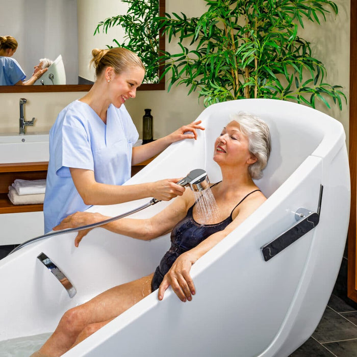 Handicare AVERO Motion Bathtub With Height-Adjustable, Wheelchair-Accessible Assisted Bathing System For Safe, Ergonomic Care Facility Use Featuring A Patient Showering With Caregiver Assist