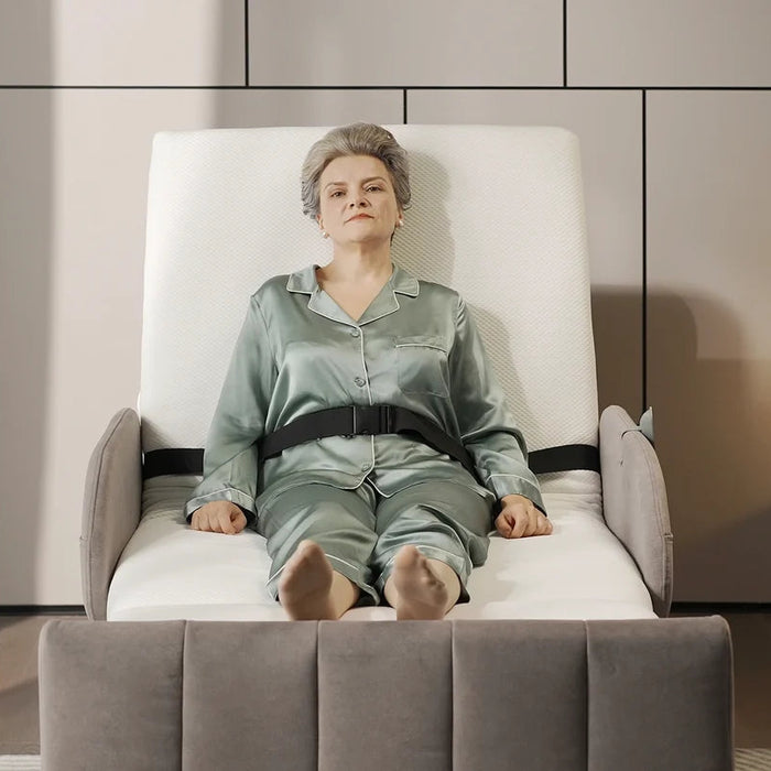 An older woman wearing a sage green pajama set sits strapped in a white and gray Starleep Orin Arlo Rotating bed chair in a modern, minimalist bedroom setting.