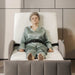 An older woman wearing a sage green pajama set sits strapped in a white and gray Starleep Orin Arlo Rotating bed chair in a modern, minimalist bedroom setting.