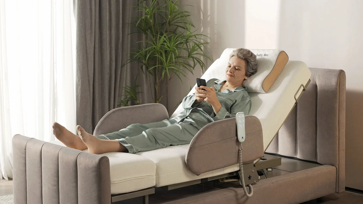 A woman in a green pajama set reclines on a beige and cream electric Arlo Orin Starsleep bed in a brightly lit bedroom, holding a smartphone.
