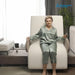 A woman wearing a sage green satin pajama set sits relaxed in a Starsleep Orin Arlo ergonomic rotating chair bed, holding the armrests. The scene is set in a modern living room with a window, curtains, and a potted plant.