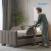 An elderly woman in a sage green pajama set pushes a bed with an attached adjustable standing and writing table frame in a modern, softly lit bedroom. The Arlo Rotating Bed logo is visible in the top right corner.