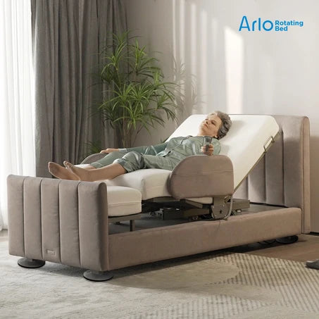 A woman reclines on a Starsleep Orin Arlo ergonomic rotating chair bed with a padded taupe upholstered frame and white mattress, shown in a softly lit bedroom setting with curtains, a plant, and a patterned rug. The Arlo Rotating Bed logo appears in the top right.