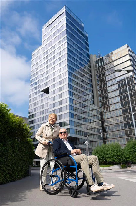 Strongback Mobility Comfort: 24+AB Wheelchair Lightweight, Ergonomic Design for Superior Comfort and Control, Modern architecture and bright, clear sky smilling elderly couple outdoors.